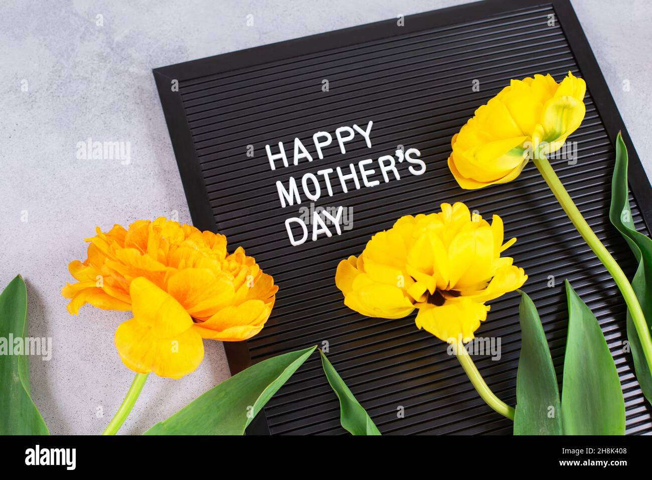 Schwarzer Karton mit weißen Plastikbuchstaben mit Zitat Happy Mothers Day und gelben Tulpen auf grauem Hintergrund. Stockfoto