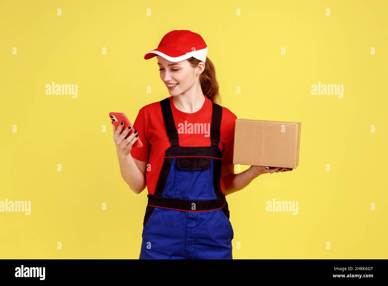 Porträt einer Kurierin, die mit dem Paket in den Händen steht und mit dem Mobiltelefon, schnellem Lieferservice, Overalls und roter Mütze trägt. Innenaufnahme des Studios isoliert auf gelbem Hintergrund. Stockfoto