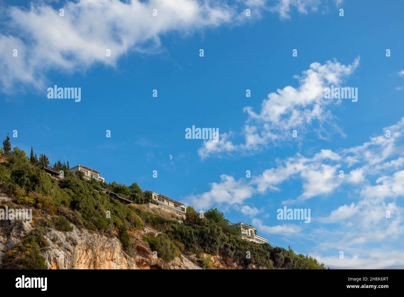 Malerische Landschaft mit griechischen Erholungsvillen auf grünen felsigen hohen Hügeln am blauen malerischen Himmel mit schönem und jungem Mond in Wolken auf der Insel Lefkada, Stockfoto