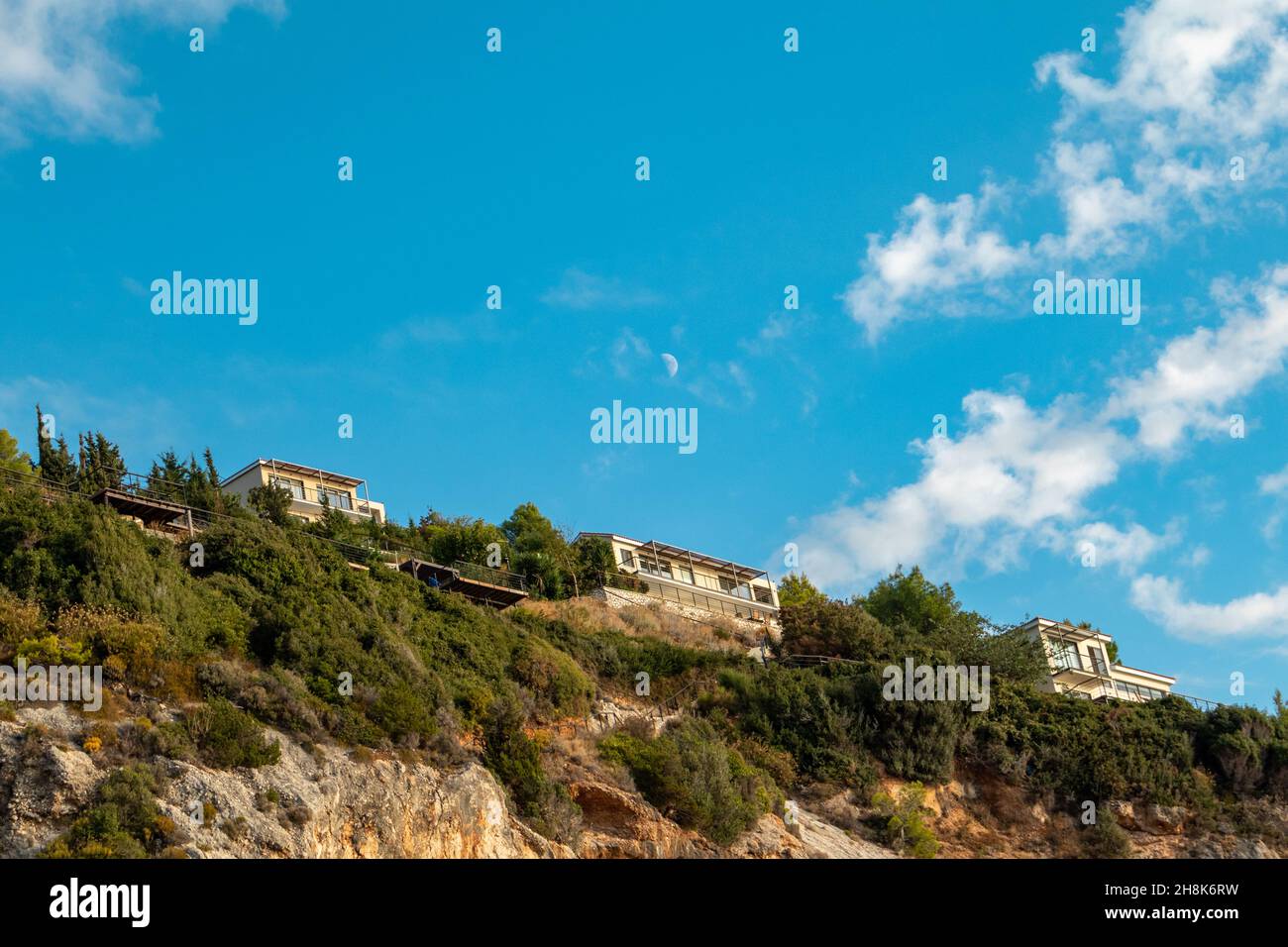 Malerische, lebhafte Landschaft mit griechischen Ferienvilla-Häusern auf grünen hohen Hügeln am blauen, malerischen Himmel mit Wolken und jungem Mond auf der Insel Lefkada, Greec Stockfoto
