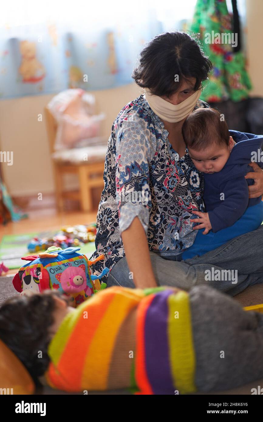 Maskierte Mutter hält ihr Baby, während sie ihr anderes Kind ansieht. Covid-19 Coronavirus Stockfoto