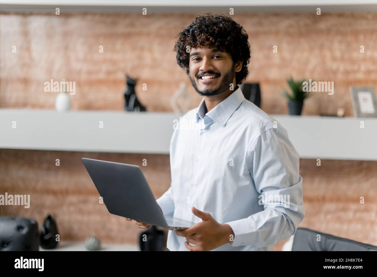 Positiv selbstbewusst moderner junger, lockiger Inder, Büroangestellter oder Freiberufler, in einem Hemd gekleidet, mit offenem Laptop im Büro stehen, mit freundlichem Lächeln auf die Kamera blicken Stockfoto