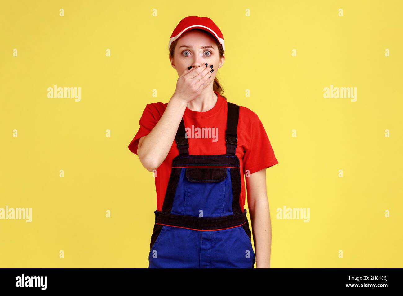 Porträt einer schockierten, staunenden Arbeiterin, die mit großen Augen auf die Kamera steht und den Mund mit einer Handfläche bedeckt, Arbeitskleidung und roter Mütze trägt. Innenaufnahme des Studios isoliert auf gelbem Hintergrund. Stockfoto