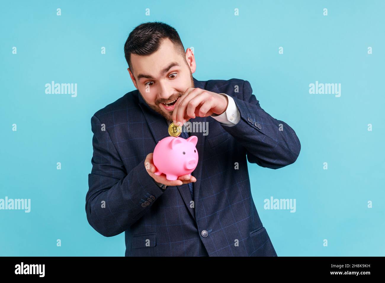 Aufgeregter Mann mit Bart, der einen Anzug im offiziellen Stil trägt, in Bitcoins investiert, goldene Krypto-Münzen ins Sparschwein gesteckt und mit Geldeinsparungen zufrieden ist. Innenaufnahme des Studios isoliert auf blauem Hintergrund. Stockfoto