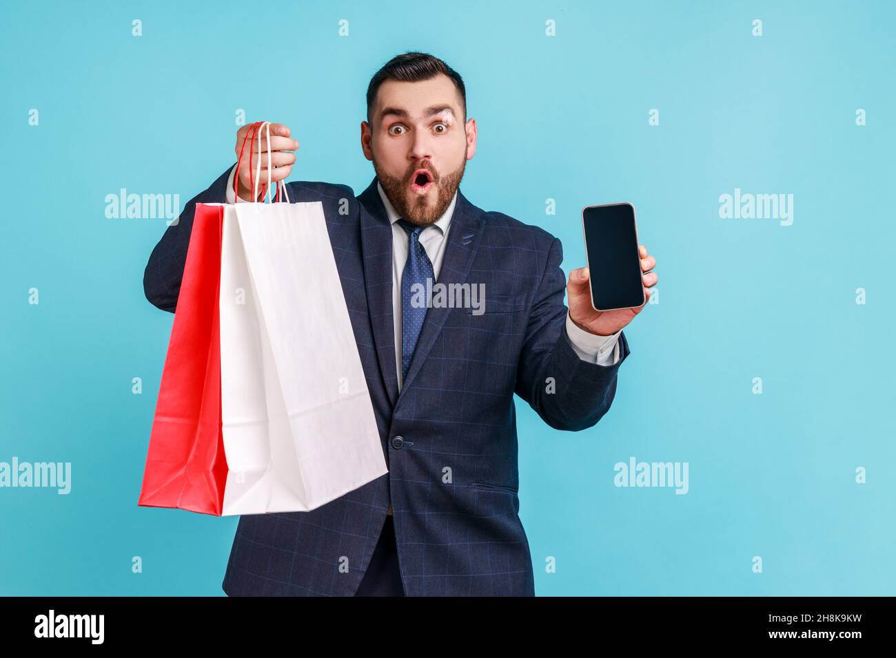 Überrascht bärtigen Mann trägt offiziellen Stil Anzug in den Händen halten Smartphone mit leerem Display und Einkaufstasche, schockiert mit einfachen Online-Shopping. Innenaufnahme des Studios isoliert auf blauem Hintergrund. Stockfoto