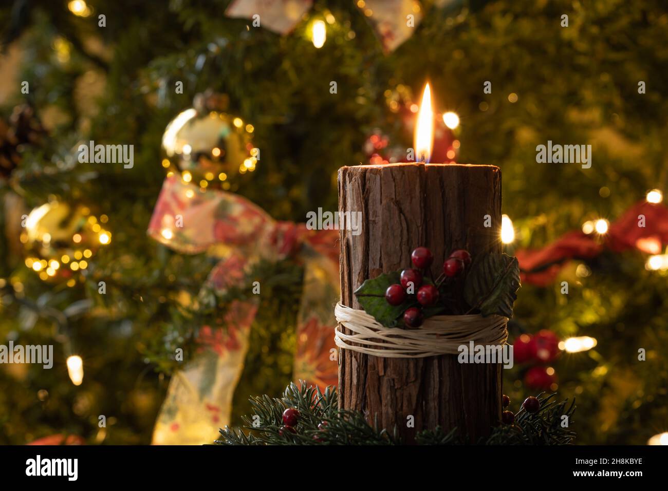 Weihnachtskerze mit roten Beeren und rustikaler Rinde, die in einem dunklen Raum vor einem geschmückten Weihnachtsbaum angezündet wird Stockfoto