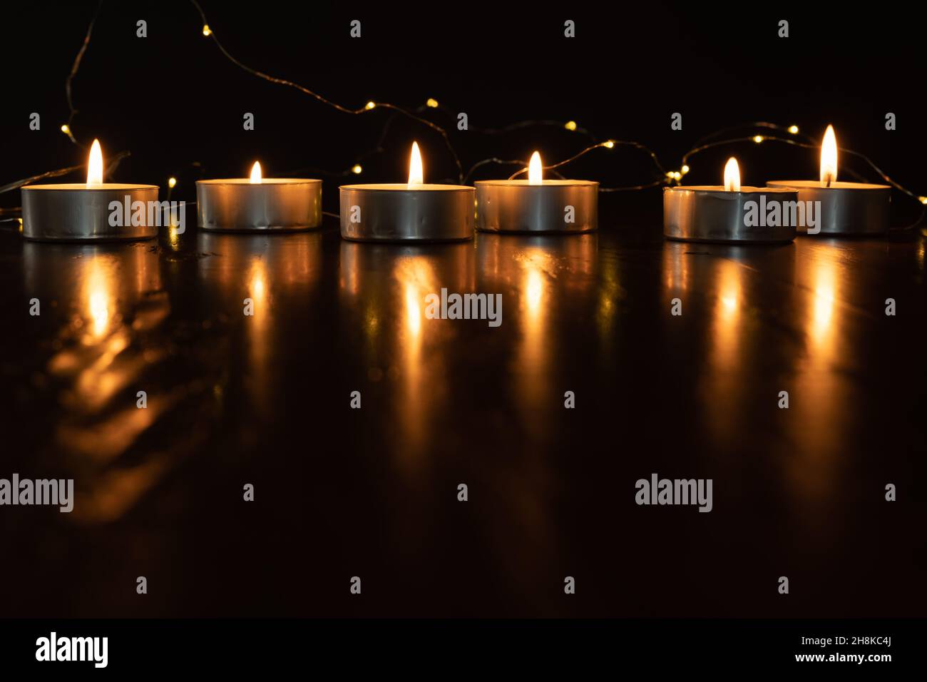 Mini-Teelicht-Kerzen brennen im Dunkeln mit Lichterketten und Flammen, die von einem dunklen Holztisch mit Platz für Kopien reflektiert werden Stockfoto