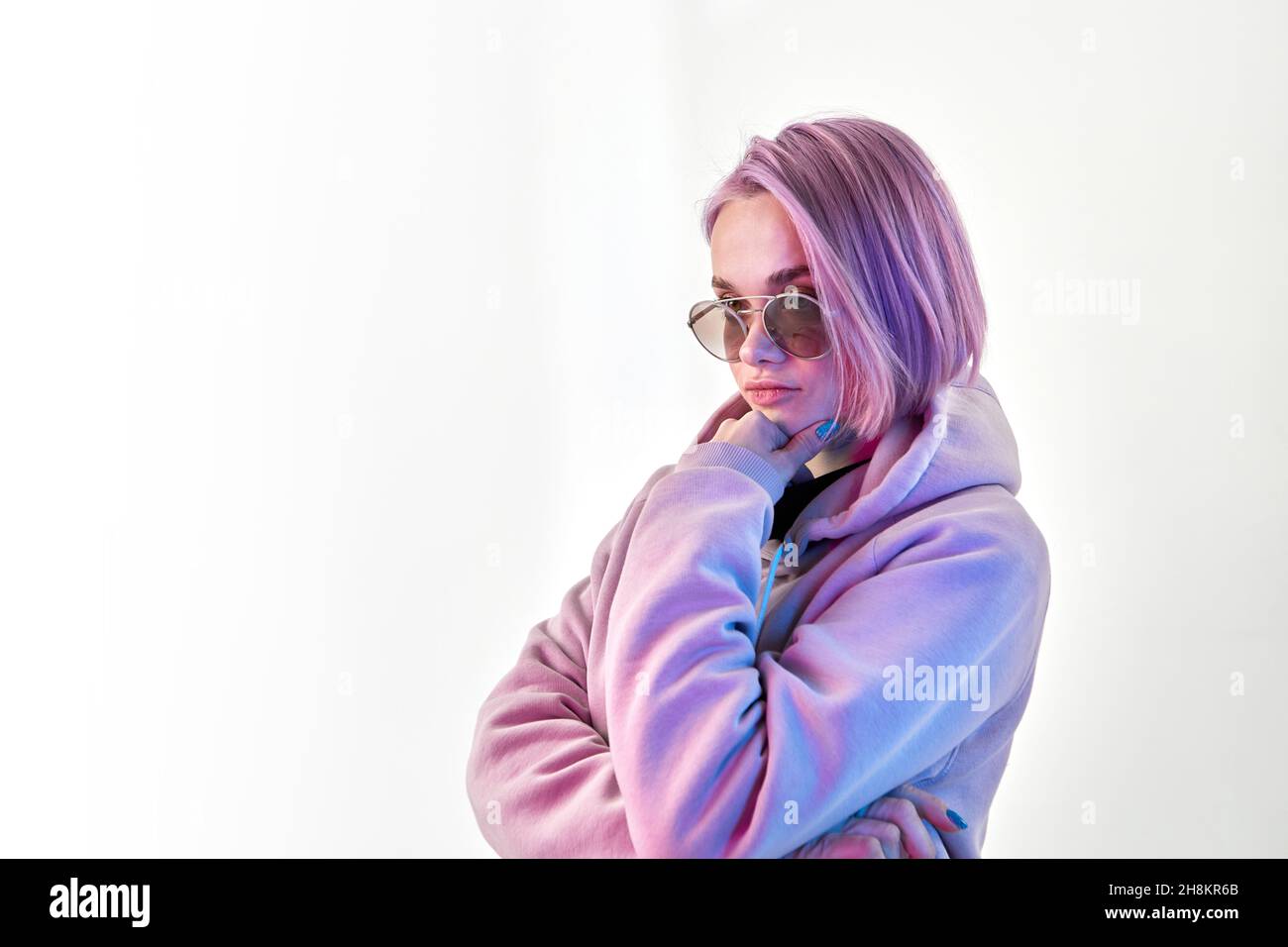 Serious junge Frau in Hoodie und stilvolle Sonnenbrille berühren Kinn und denken über die Entscheidung auf weißem Hintergrund Stockfoto