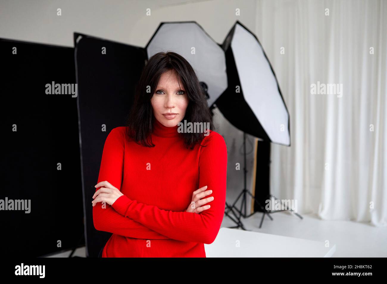 Seriöse Frau im roten Rollkragen, die mit gefalteten Händen im Studio mit Ausrüstung für Fotoaufnahmen steht und die Kamera anschaut Stockfoto