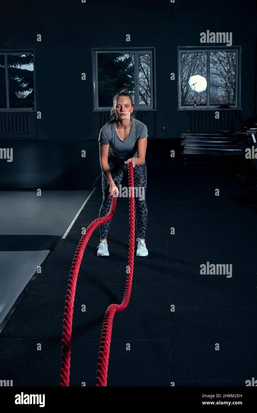 Vertikale Ganzkörperaufnahme einer athletischen Fitness-Frau, die im CrossFit-Fitness-Studio ein funktionelles Training mit Kampfseilen macht Stockfoto