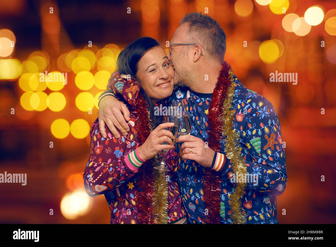 Fröhliches romantisches Paar mittleren Alters, das zu Weihnachten küsst und Champagner trinkt, um die Weihnachtszeit zu feiern, gekleidet in farbenfrohe Weihnachten-Themen-Tops ag Stockfoto