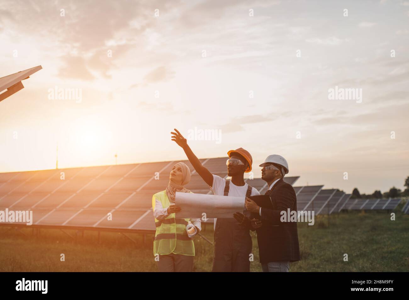 afroamerikanische Technikerin im Gespräch mit muslimischer Frau und indischem Mann während der Arbeitssitzung auf einer Solarstation. Industriearbeiter diskutieren gemeinsames Projekt der grünen Energieerzeugung. Stockfoto