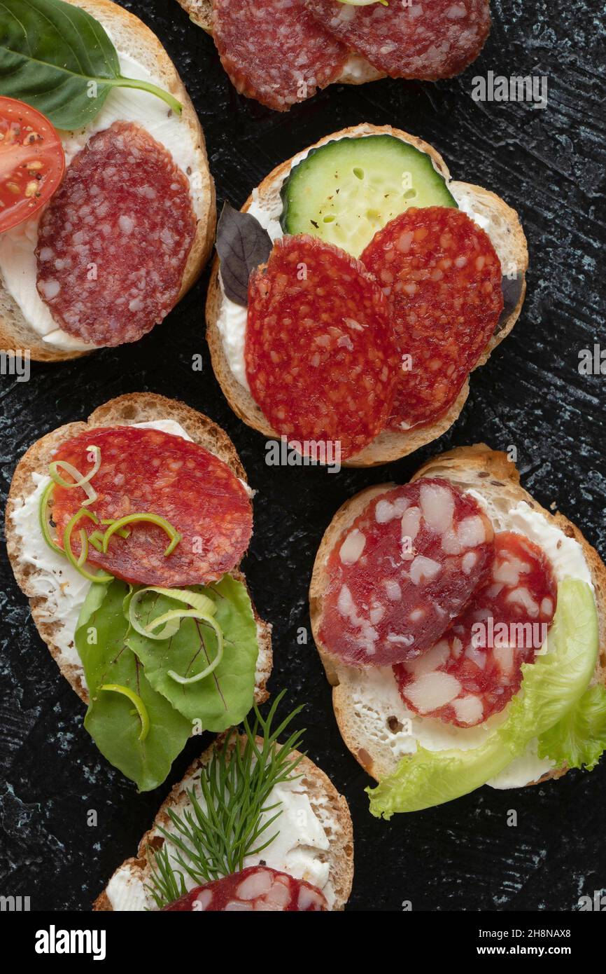 Vertikales Foto mit Nahaufnahmen von Sandwiches oder Snacks mit verschiedenen Wurst- und Gemüsesorten auf schwarzem Hintergrund. Weichfokus. Stockfoto