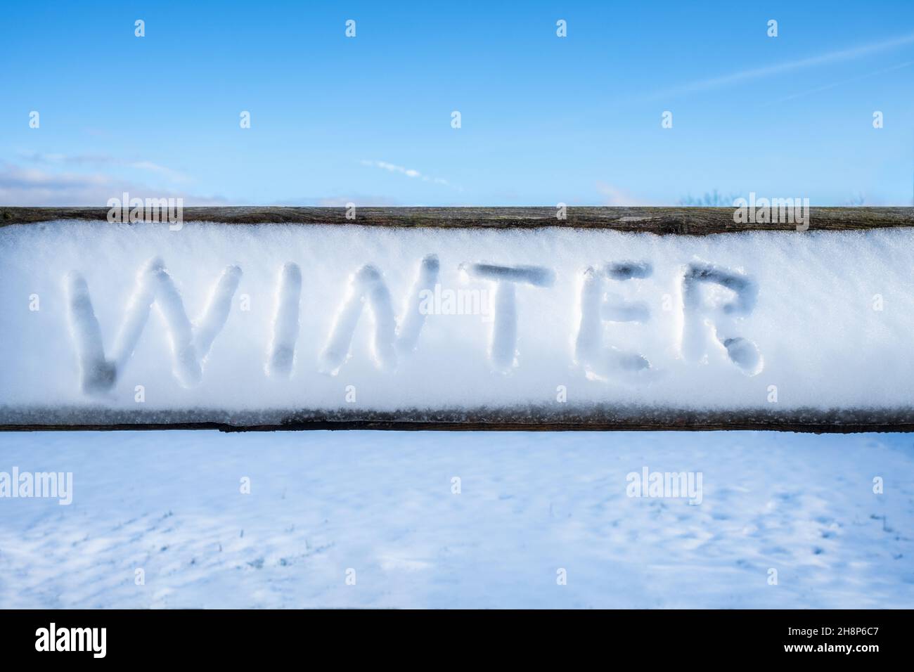Das Wort „Winter“ wurde an einem sonnigen Wintertag im Schnee auf einer Holzdiele handgeschrieben. Unberührtes schneebedecktes Feld und blauer Himmel im Hintergrund. Winterkonzept Stockfoto