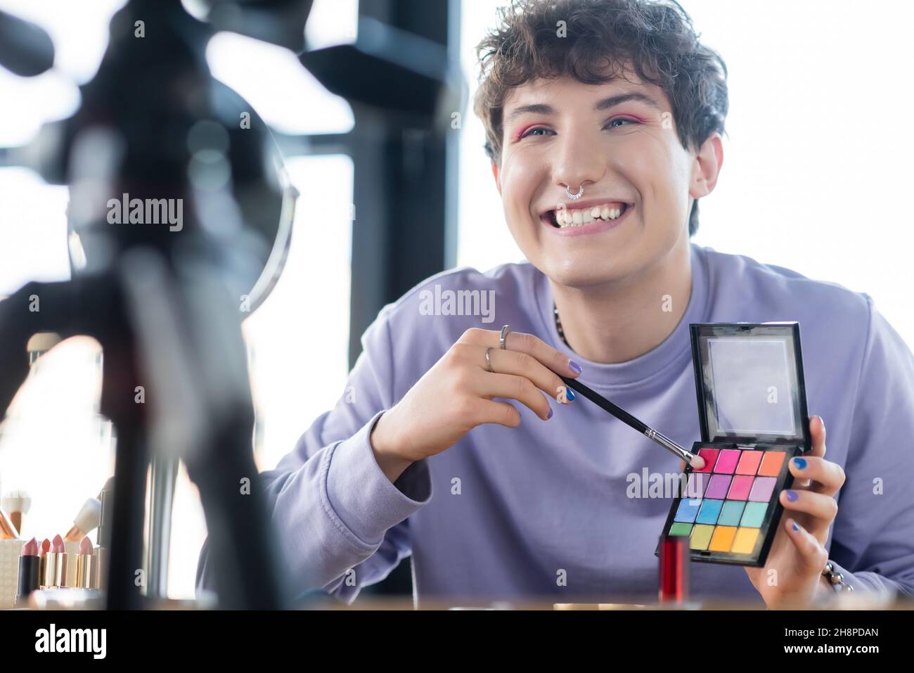 Lächelnde Transgender-Person, die Augenschatten und kosmetische Bürste in der Nähe von Stativ und Spiegel hält, Stockbild Stockfoto