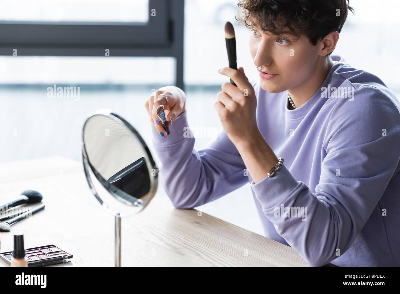 Junge Transgender Make-up-Künstler hält kosmetische Pinsel in der Nähe des Spiegels, Stock Bild Stockfoto