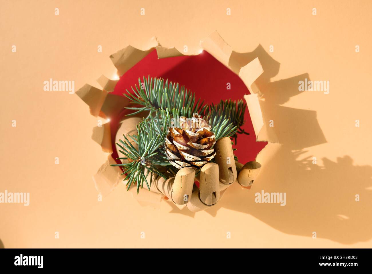 Frohe Weihnachten, Hand mit Tannenzweigen und Kegel durch Papierloch. Kreativer minimalistischer Konzepthintergrund mit beigefarbenem, orangefarbenem Papier. Stockfoto