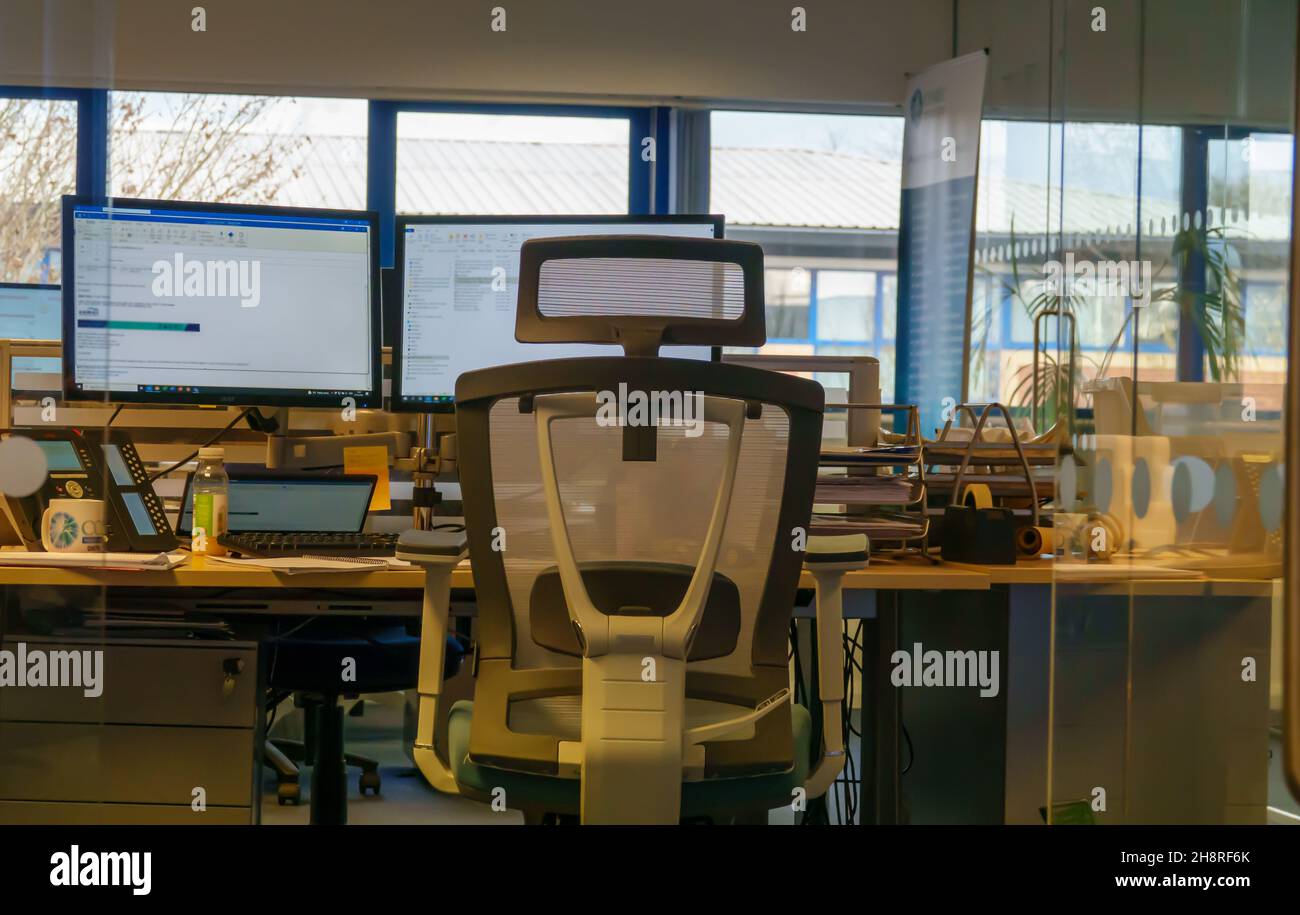 Blick auf die Rückseite eines leeren ergonomischen Bürostuhls vor einem geschäftigen Schreibtisch und auf zwei Computerbildschirme in einem Großraumbüro Stockfoto