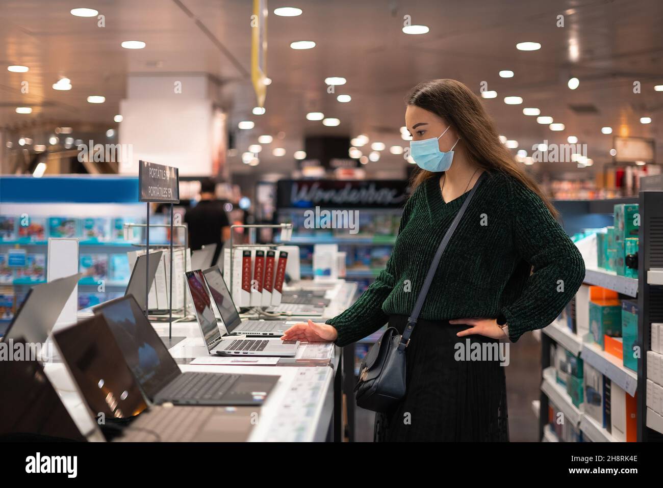 Weibliche Kundin in Maske hält Hand an der Taille und untersucht modernes Netbook, während sie während einer Pandemie das Einkaufszentrum besucht Stockfoto