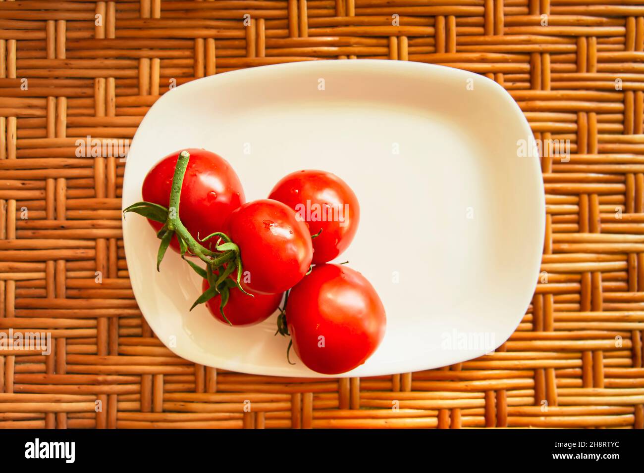 Eine Gruppe von roten Kirschtomaten auf einer weißen flachen rechteckigen Platte stehen auf einer hölzernen Korboberfläche. Blick von oben. Horizontales Foto. Lifestyle-Komposition. Nahaufnahme. Hochwertige Fotos Stockfoto