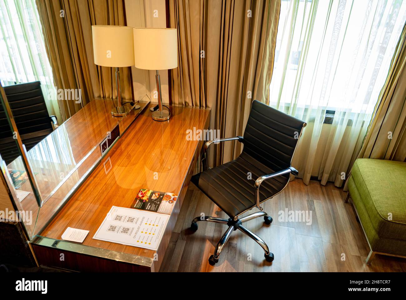 Ein moderner Arbeitsbereich mit einem eleganten Holztisch, einer Tischlampe, einem großen Spiegel und einem schwarzen Sessel in einem Deluxe-Zimmer. Stockfoto