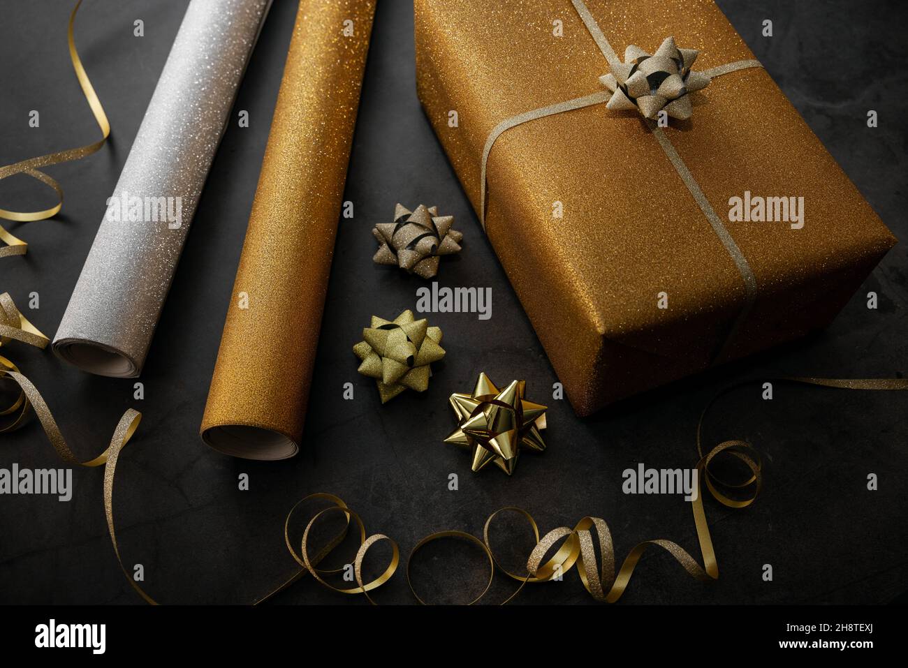 Geschenkverpackung. Eingewickeltes goldenes Geschenk mit Geschenkpapierrollen und Accessoires auf schwarzem Stein Hintergrund Stockfoto