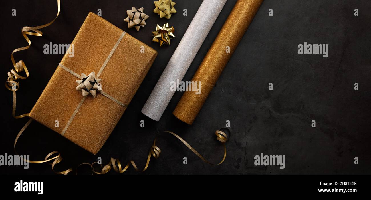 Geschenkverpackung Service. Eingewickelte goldene Geschenkbox mit Geschenkpapierrollen auf dunklem Stein Hintergrund. Banner Copy Space Stockfoto