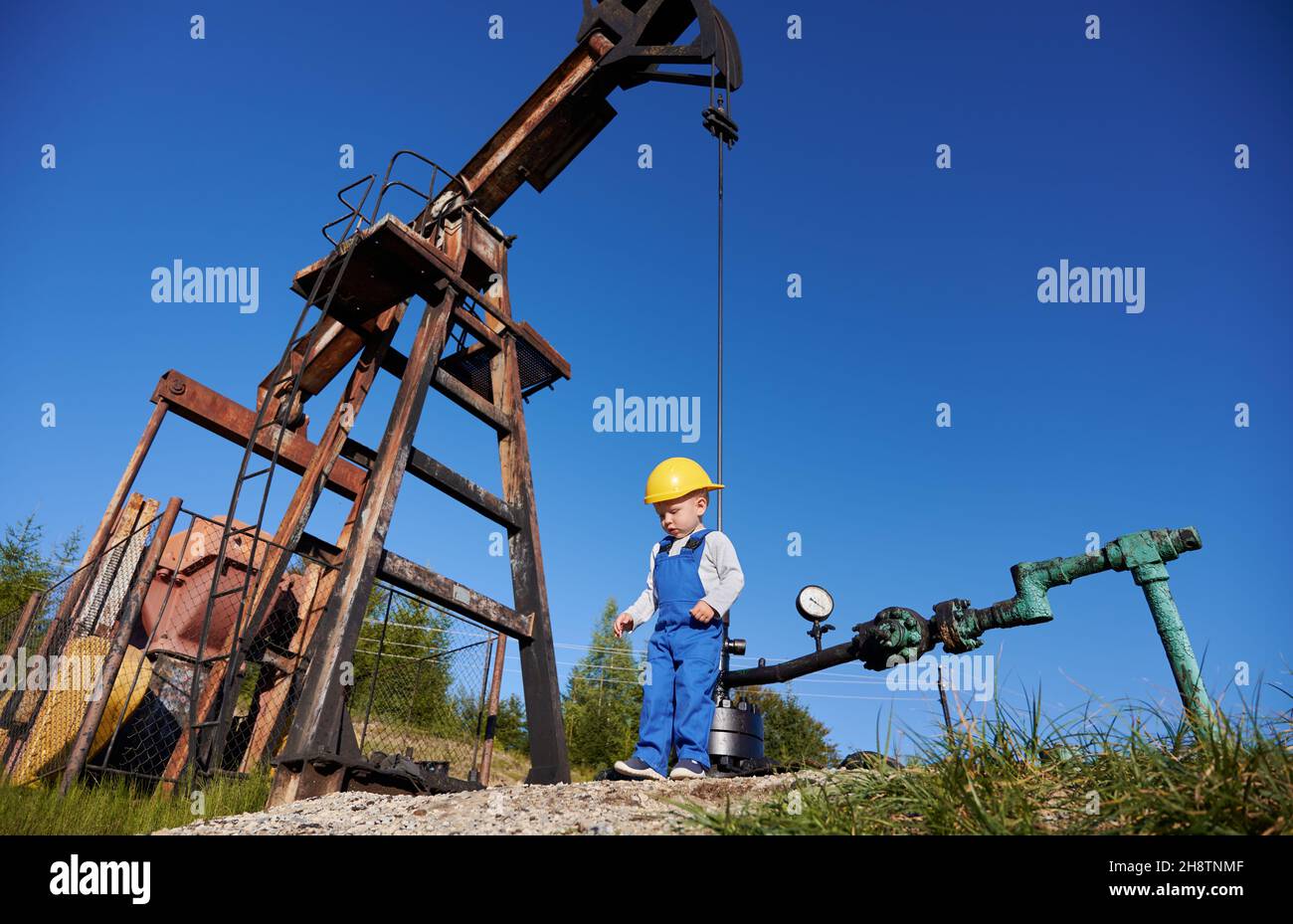 Ansicht von unten eines kleinen jungen Energietechnikers in der Nähe der Ölplattform. Boy in Overalls gegen den blauen Himmel, Petroleum-Pipeline und Pumpenheber für die Gewinnung von natürlichem Kraftstoff. Stockfoto