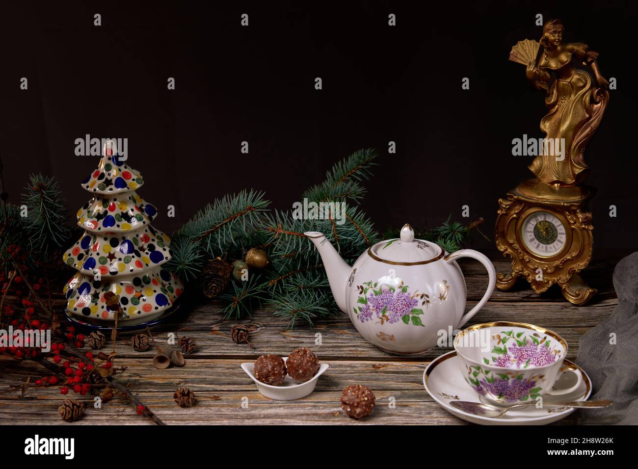 Weihnachts-Teekannen und -Teetassen, Süßigkeiten in einer Keramikplatte, Christbaumzweige, Keramik-Weihnachtsbaum und eine antike Uhr auf einem Holztisch. Stockfoto