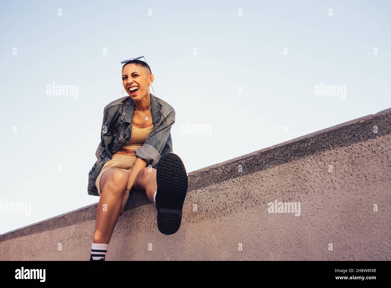 Fröhliche junge Frau, die Spaß hat, während sie draußen an einer Wand sitzt. Fröhliche junge Frau, die lacht und sich amüsiert. Junge Hipster Frau Gefühl Stockfoto