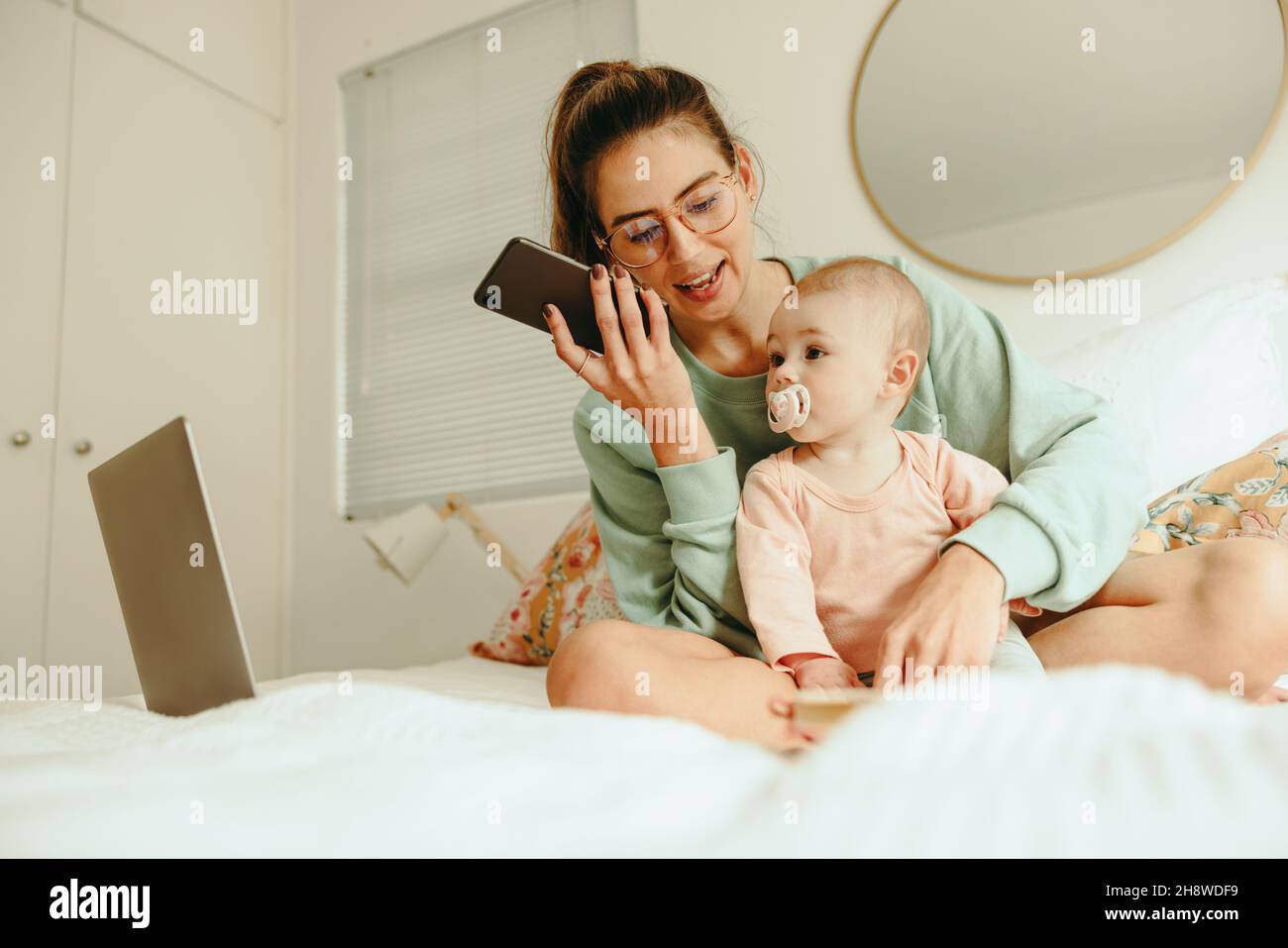 Multitasking-Mutter, die einen Anruf entgegennimmt, während sie mit ihrem Baby auf einem Bett sitzt. Alleinerziehende Mutter plant mit ihren Kunden, während sie von zu Hause aus arbeitet. Neue Mo Stockfoto