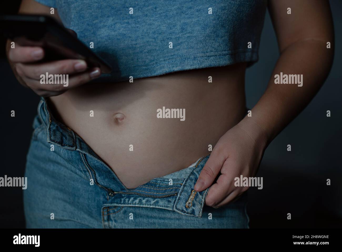 Frauenbauch in unknöpfbarer blauer Jeans. Weibliche Figur. Eine Frau hält ein Telefon in den Händen. Stockfoto