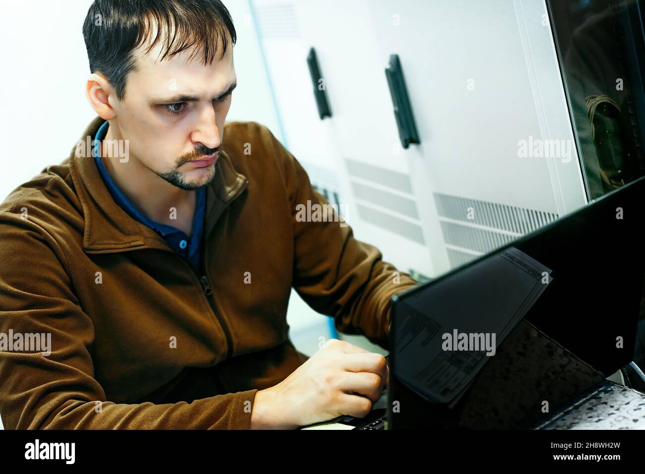 Porträt eines Programmierers bei der Arbeit. Eine authentische Szene. Ein IT-Spezialist arbeitet seit 30-40 Jahren an einem Laptop im Serverraum. Führen Sie Diagnosen aus oder führen Sie Wartungsarbeiten durch. Hintergrund. Stockfoto
