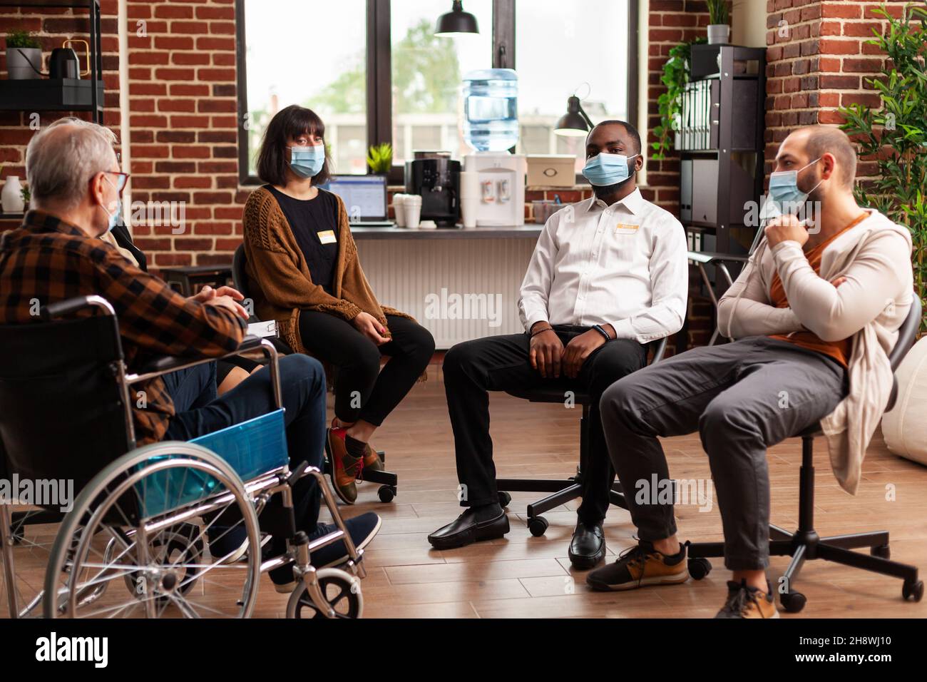 Multiethnische Personen, die während einer Coronavirus-Pandemie an einer Gruppentherapie teilnehmen. Alter Mann sitzt im Rollstuhl und Patienten im Kreis Vorbereitung auf ein Treffen Diskussion und Beratung. Stockfoto
