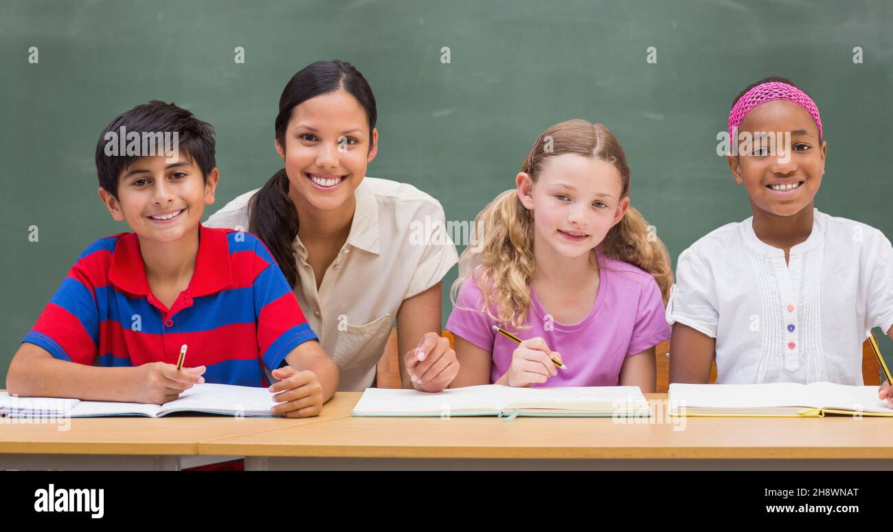 Porträt eines lächelnden Lehrers mit einem Schuljungen und Schülerinnen am Schreibtisch gegen eine Tafel im Klassenzimmer Stockfoto