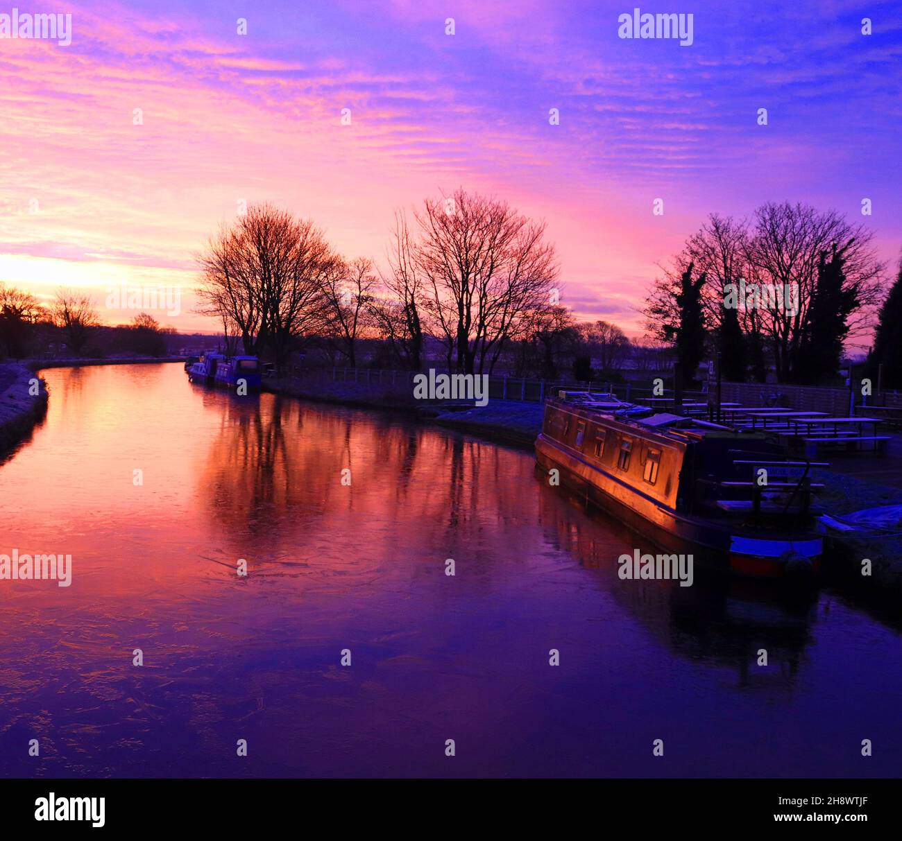 Die rote, feuerrote Sonne kommt gerade über einem blauen, eisigen und kalten Kanal von Leeds und Liverpool auf, an einem Herbstmorgen, der ein Narrowboat mit einem orangefarbenen Leuchten beleuchtet Stockfoto