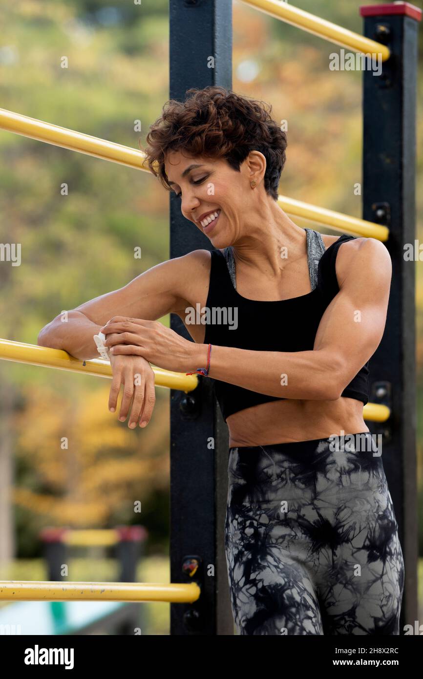 Selbstbewusste, muskulöse hispanische Läuferin mit kurzen Haaren im aktiven Ohr, die Benachrichtigungen auf der Smartwatch während des Outdoor-Trainings im Park auf der Sonne überprüft Stockfoto