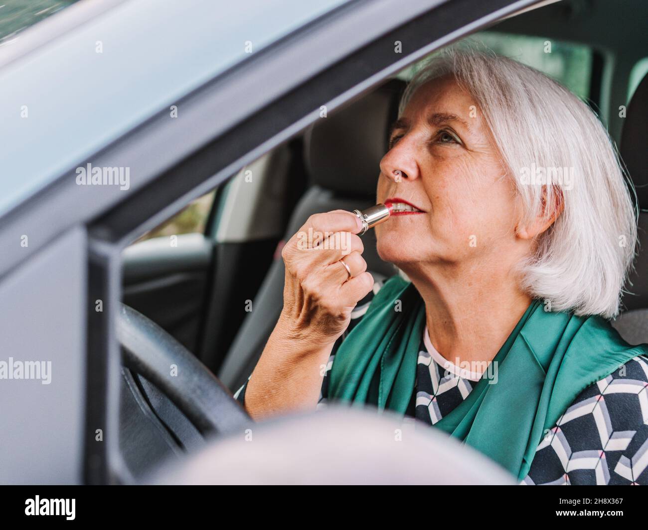 Ältere Frau in eleganter Kleidung, die Lippenstift in den Rückspiegel trägt, während sie auf dem Fahrersitz eines modernen Autos sitzt Stockfoto