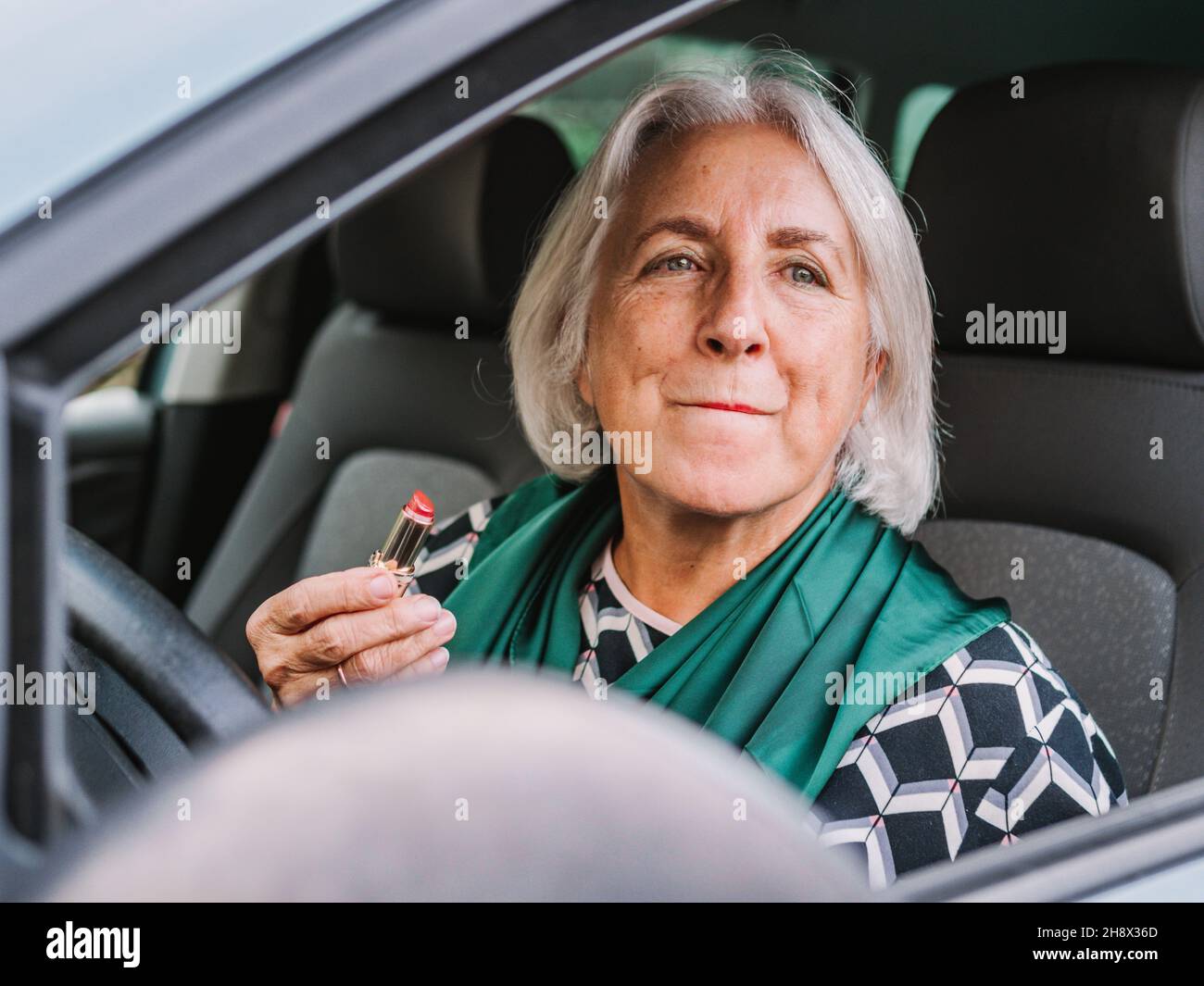 Ältere Frau in eleganter Kleidung, die Lippenstift aufträgt, während sie auf dem Fahrersitz eines modernen Autos sitzt Stockfoto