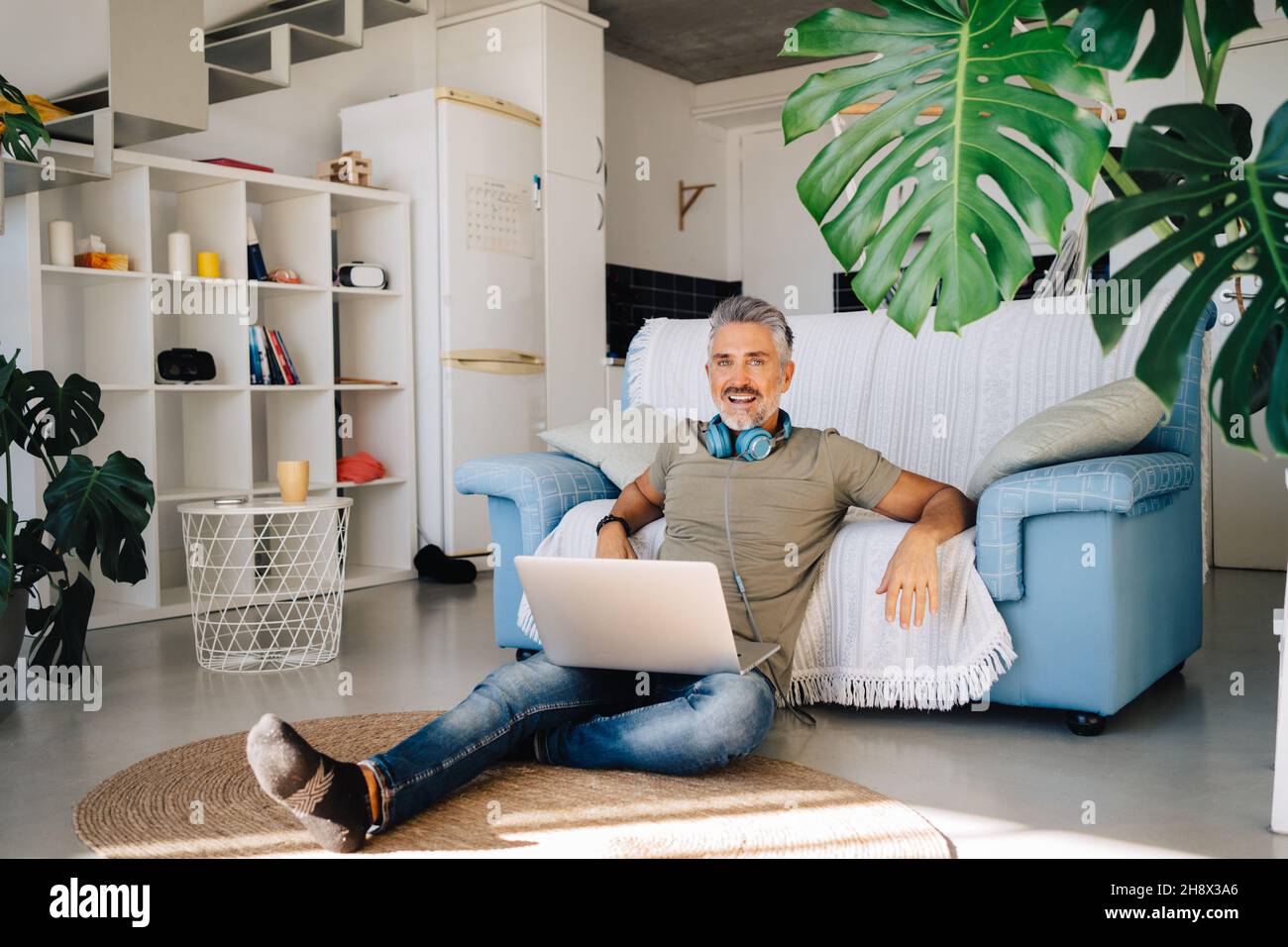 Grauhaariger, glücklicher, gutaussehender Mann, der Laptop und Kopfhörer zum Musikhören benutzte, während er sich in der Nähe einer bequemen Couch im Loft-Apartment abkühlte Stockfoto