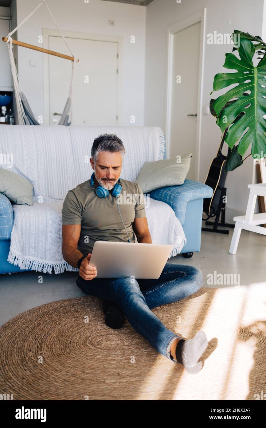 Grauhaariger, hübscher Mann, der Laptop und Kopfhörer zum Musikhören benutzt, während er sich in der Nähe einer bequemen Couch im Loft-Apartment abkühlte Stockfoto