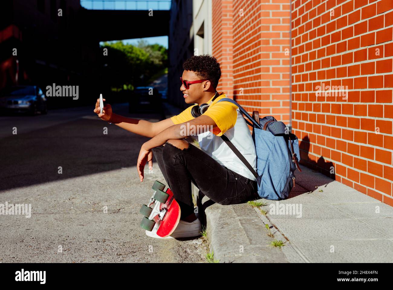 Junger schwarzer Mann mit Sonnenbrille sitzt auf der Straße und porträtiert sich selbst mit dem Smartphone Stockfoto