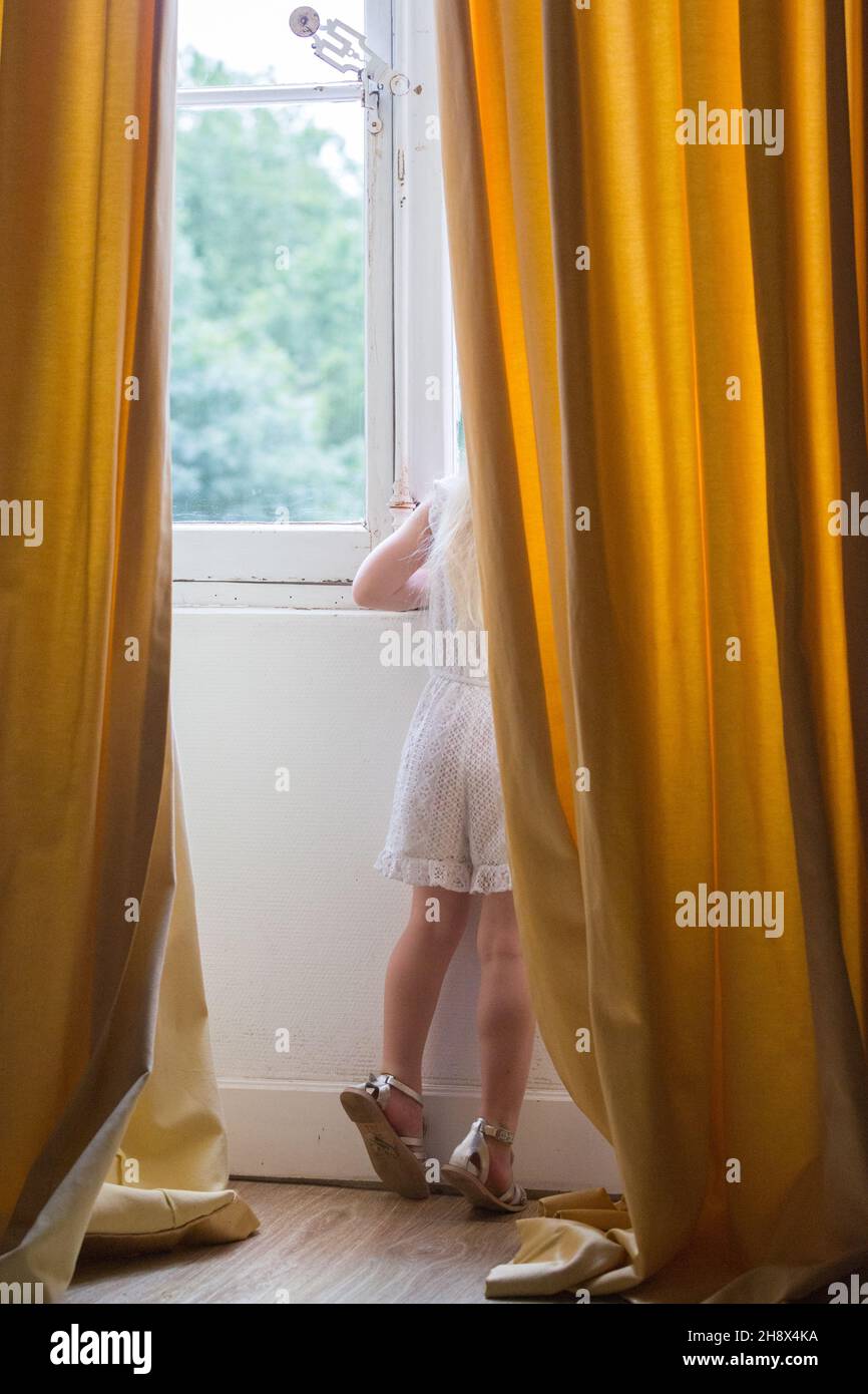 Kleines Mädchen in weißer Kleidung, das hinter einem Vorhang in einem französischen Schloss aus einem Fenster spähend Stockfoto