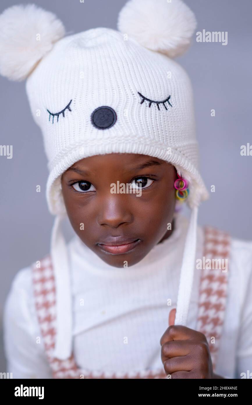 Entzückende afroamerikanische Mädchen Modell in stilvollen warmen Hut mit Pompoms Blick auf die Kamera vor grauem Hintergrund Stockfoto