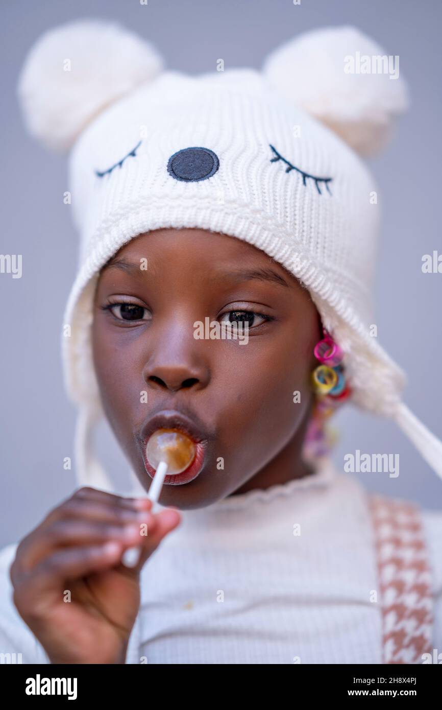 Liebenswert kleine afroamerikanische Mädchen in warmen Hut leckt süßen Lollipop und Blick weg vor grauem Hintergrund Stockfoto
