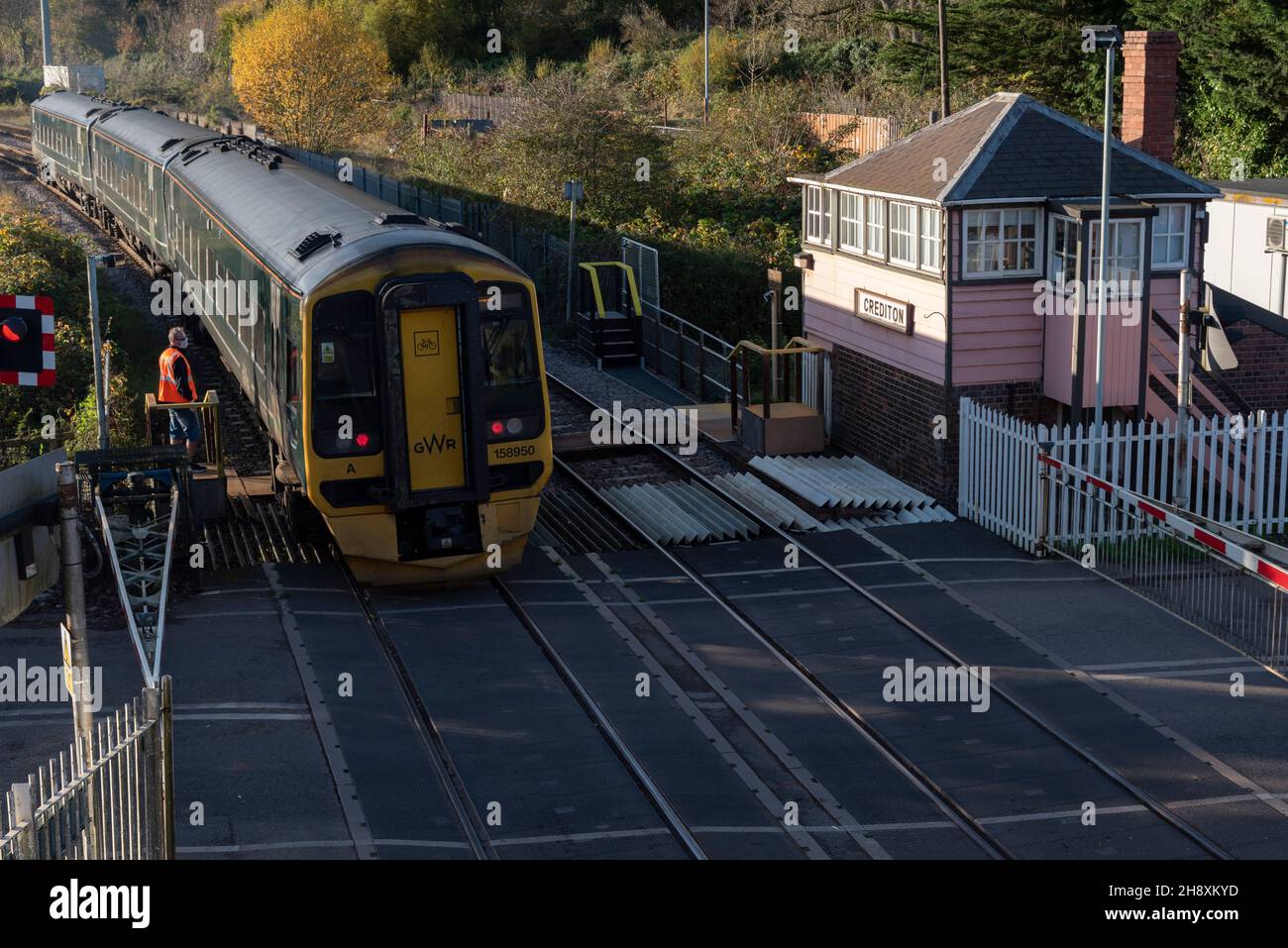 Crediton, Devon, England, Großbritannien. 2021. Passenegr-Zug fährt vom Bahnhof Crediton und dem Bahnübergang ab. Beobachtet von einem Signalmann, der einen Schlüssel für Th ausgibt Stockfoto