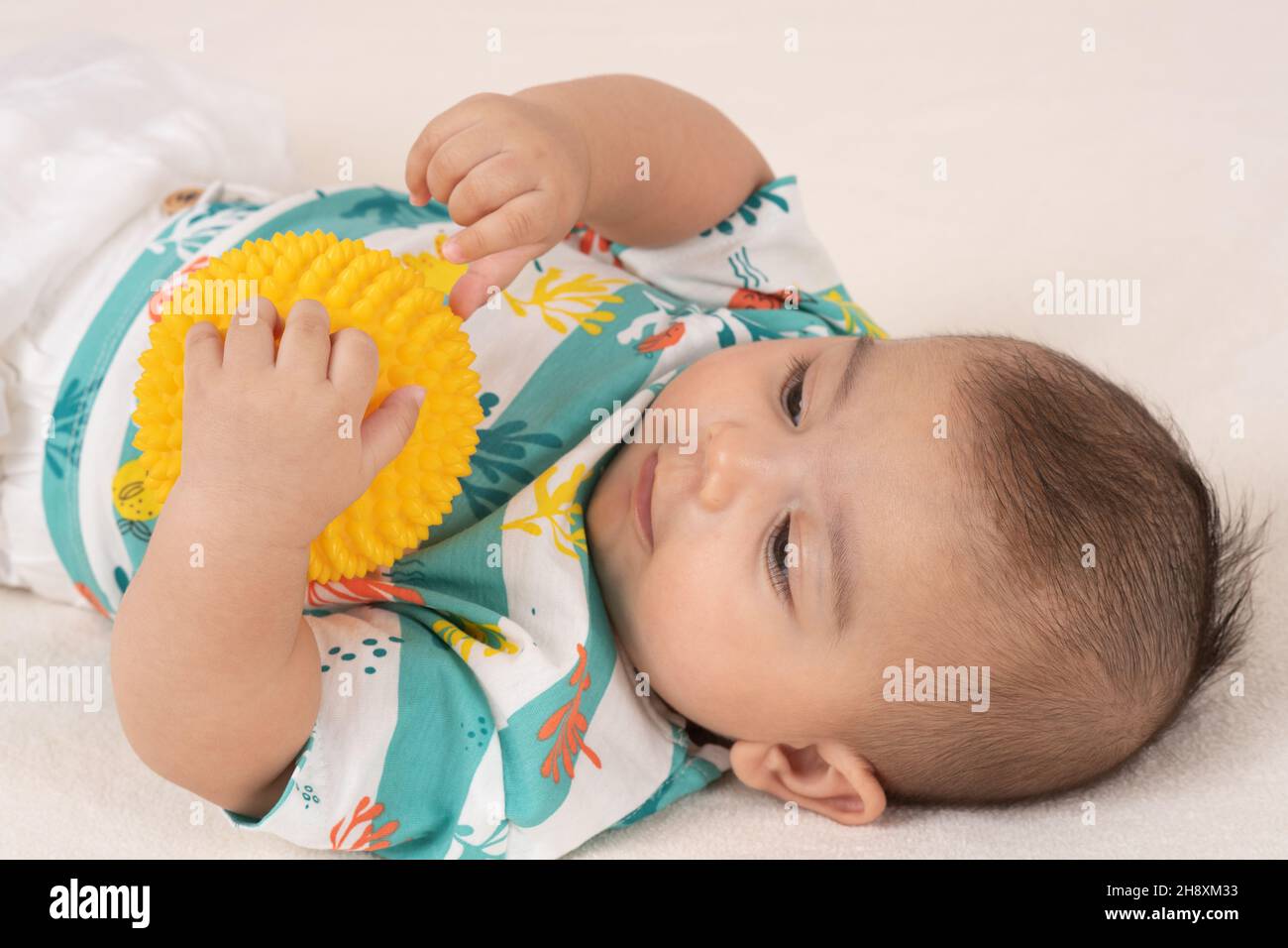 3 Monate alter Junge auf dem Rücken hält Plastikspielzeugkugel mit einer Hand und fühlt Textur von Noppen mit der anderen Hand, mit Fingern Stockfoto