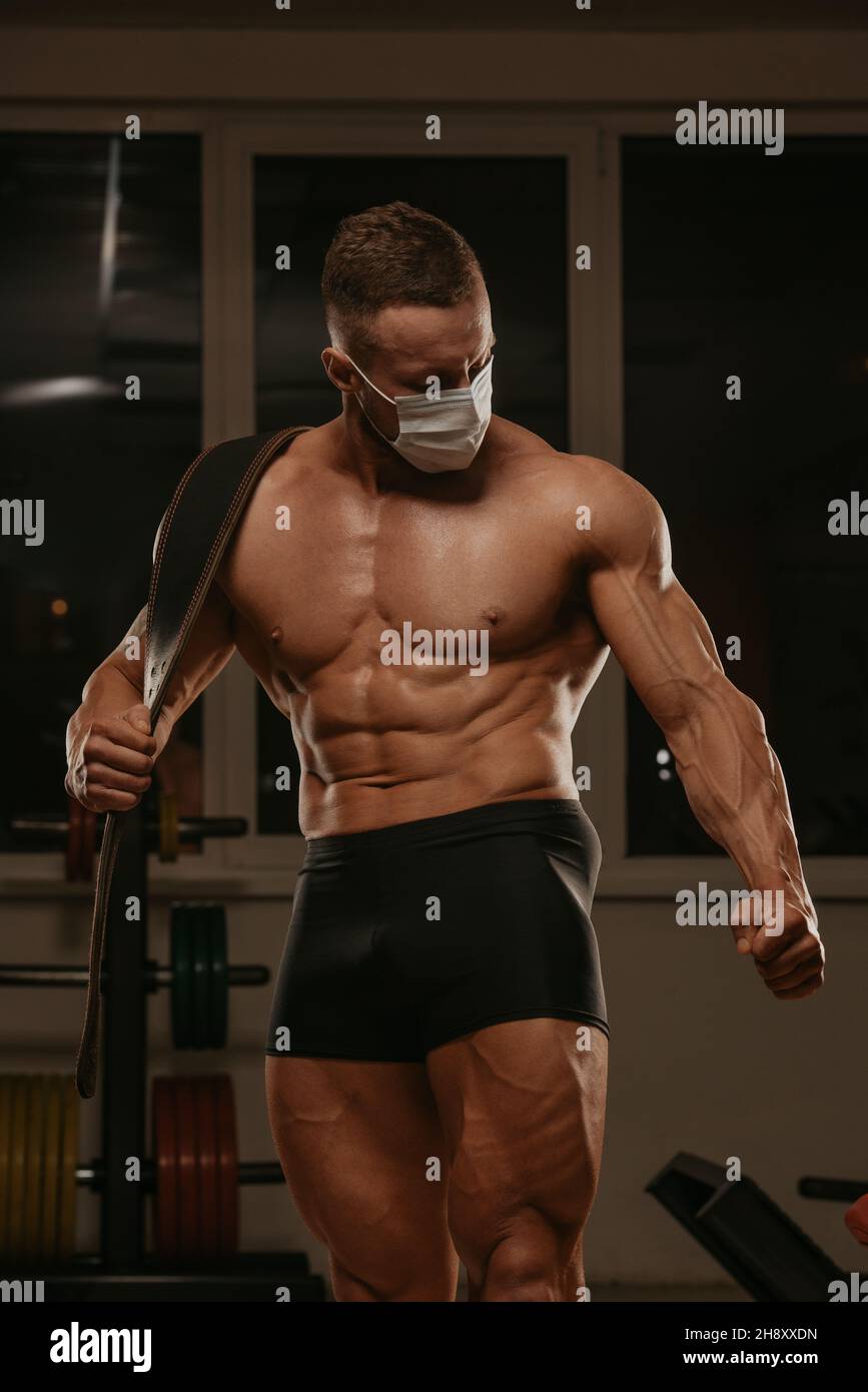 Eine mittellange Aufnahme eines gerafften Bodybuilders in einer medizinischen Gesichtsmaske, der nach einem Training posiert. Ein muskulöser Kerl in einer OP-Maske spannt seinen m an Stockfoto