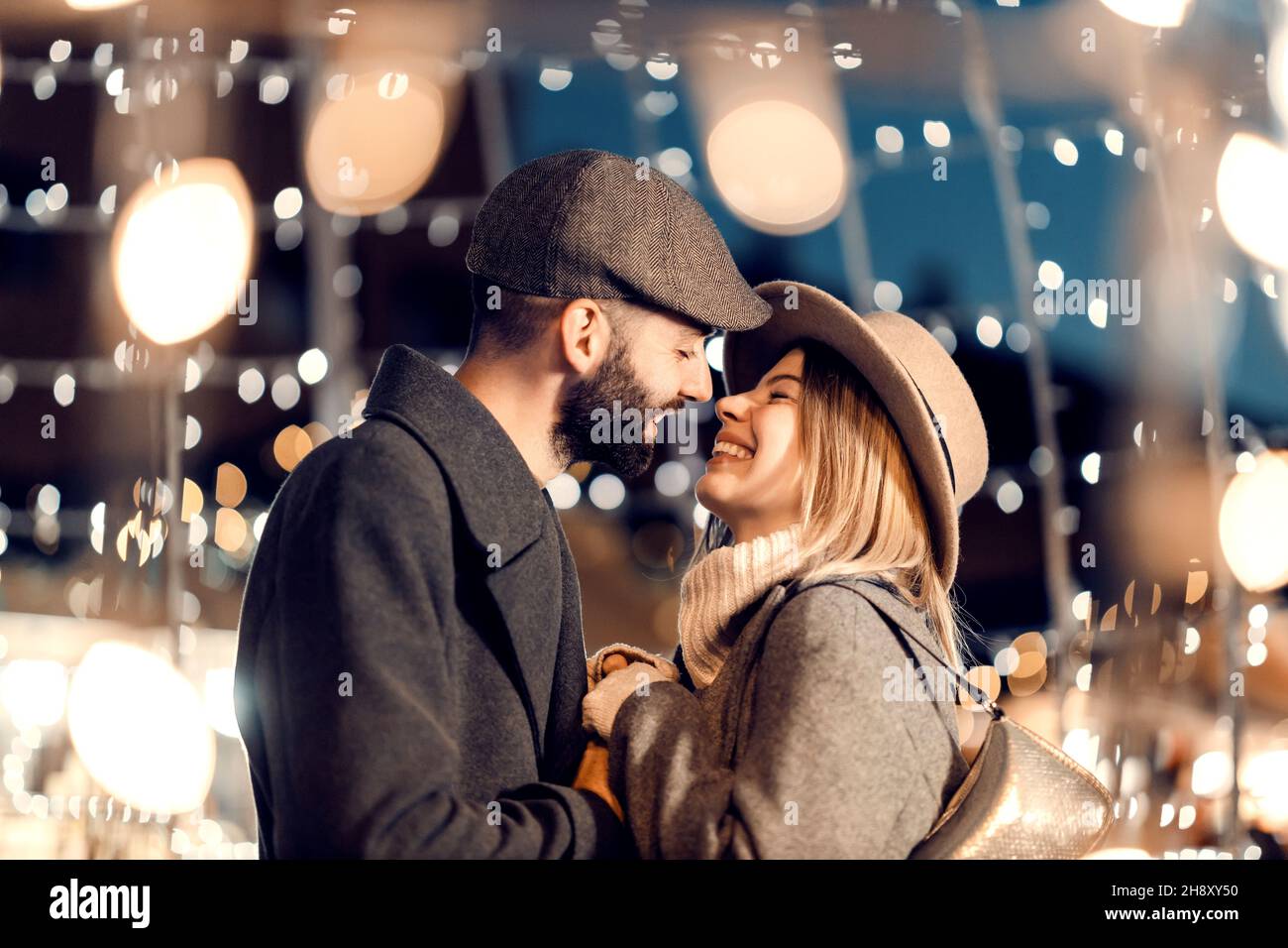 Weihnachtsliebhaber küssen sich am Silvesterabend im Freien. Ein junges, glückliches Paar steht auf der Straße, umgeben von Lichtern, und küsst sich am Silvesterabend Stockfoto