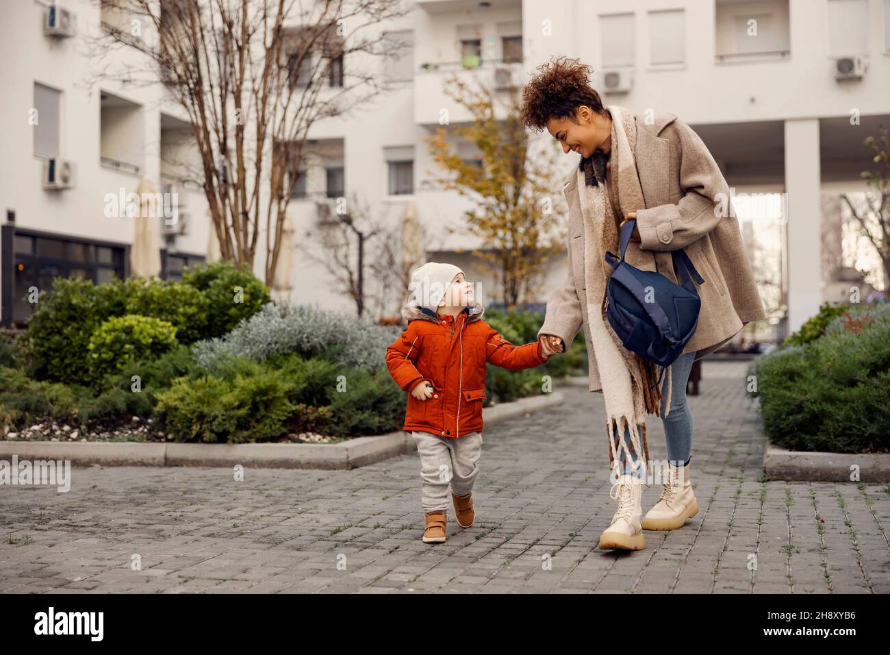 Pfleger, der den Kindergartenjungen in den Kindergarten bringt. Die Betreuerin trägt einen kleinen Rucksack und bringt den kleinen Jungen in den Kindergarten. Stockfoto